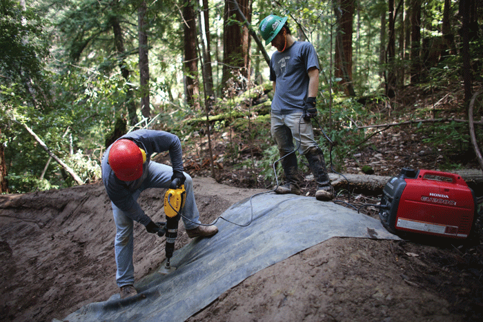 MBoSC Trail Construction Manager Drew Perkins looks on as a volunteer compacts a berm with a demolition hammer.