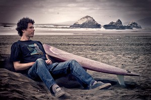 Photo of Jaimal Yogis sitting on the beach next to his surfboard. 