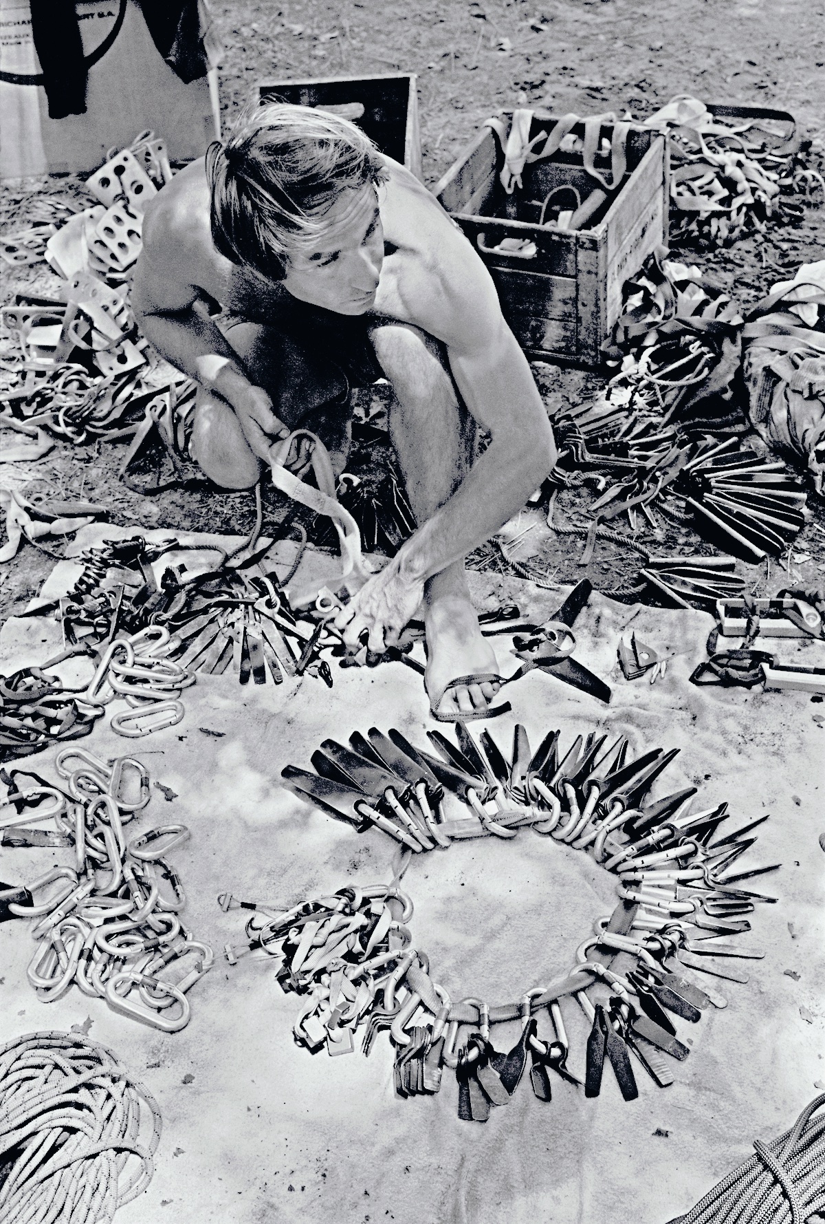 Black and white photo Yvon Chouinard squatting with an organized circle of climbing pitons in front of him