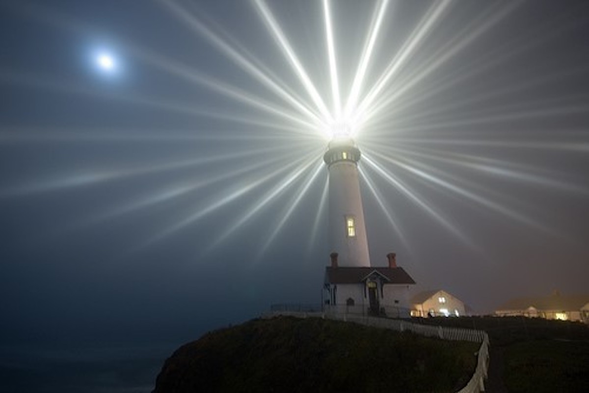 A photo of Pigeon Point Lighthouse at night with beams of light radiating from the lighthouse